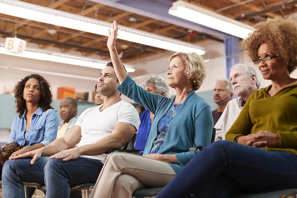 A diverse group of adults sitting in chairs at a community meeting, with one middle-aged woman raising her hand to speak to her homeowners association management company.