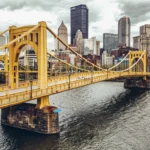 Yellow bridge over Pennsylvania river, Pittsburgh skyscrapers in background.